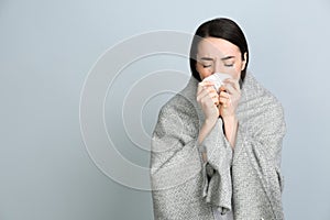 Young woman with blanket suffering from runny nose on light grey background. Space for text