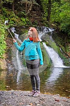 Young woman blogger in autumn mountains near lake and waterfall is recording video, taking photo with selfie stick