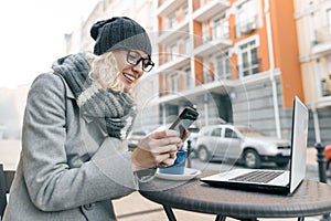 Young woman blogger freelancer in warm clothes hat in outdoor cafe with computer laptop, mobile phone, city street background