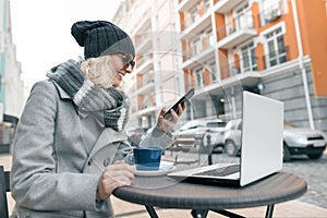 Young woman blogger freelancer in warm clothes hat in outdoor cafe with computer laptop, mobile phone. Urban background