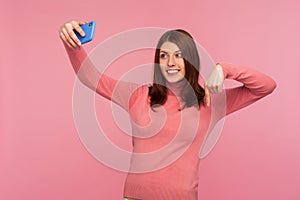 Young woman blogger in pink sweater recording video message to her admirers on smartphone asking to subscribe and rate her posts