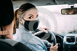 A young woman with blond hair in a black medical mask and a turquoise jacket is driving a left-hand drive