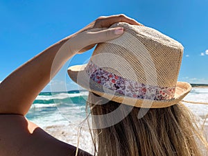 Young woman with blond hair and hat staring at the sea and sky from the beach in a windy summer day