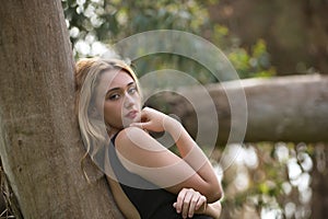Young woman, blonde and beautiful, with a black dress, leaning on the trunk of a tree, in the middle of nature, pure and virginal