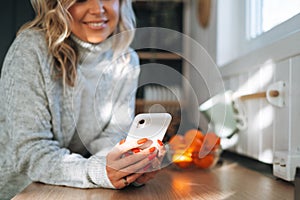 Young woman with blonde curly hair in grey sweater using mobile phone in hands in kitchen at house