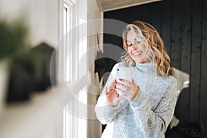 Young woman with blonde curly hair in grey sweater using mobile phone in hands near window at house