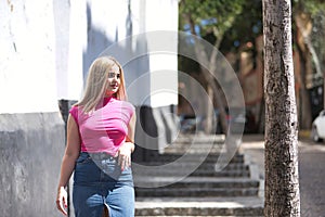 Young woman, blonde, green eyes, wearing pink t-shirt and denim skirt, walking down one of the streets of the city, going down
