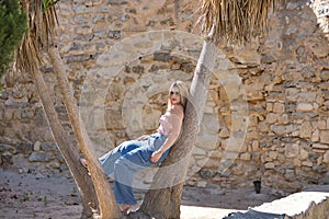 Young woman, blonde, green eyes, wearing pink top and jeans, leaning against the trunks of a palm tree, with a serious and defiant