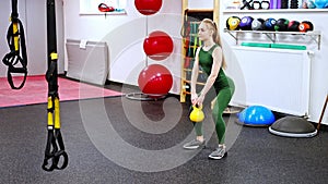 Young woman with blonde hair squatting with kettlebells in gym