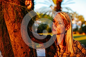 Young woman with blonde long hair standing near palm tree outdoors looking pensive in sunset