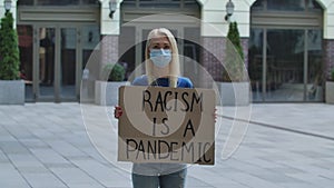 Young woman blonde in medical mask stands with a cardboard poster RACISM IS A PANDEMIC in a public place outdoor. Single