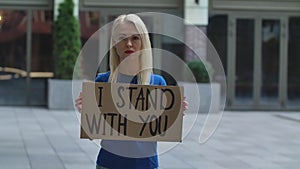 Young woman blonde stands with a cardboard poster I STAND WITH YOU in a public place outdoor. Single protest. No racism