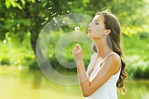 Young woman blowing dandelion in meadow. Beautiful female makes a wish on sunny summer day in park. Outdoors. Enjoy nature.
