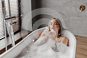 Young woman blowing soap bubbles and having fun while lying in bathtub full of foam at home, copy space