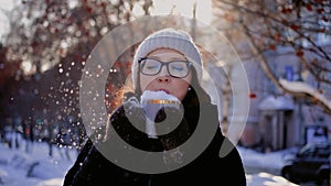 A young woman blows snow off a coffee mug on a snowy city street on a winter day