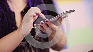 A young woman with blue Afrokos at home on the couch at a table with a laptop is texting in a smartphone in instant messengers