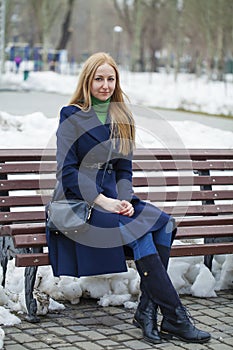 Young woman in a blue coat sitting on a bench in winter park