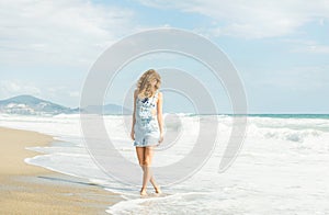 Young woman in blue denim jumpsuit walking along beach and the stormy ocean on sunny day