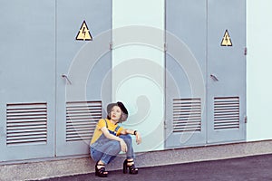 Young woman in blue denim overalls and yellow tshirt with black hat sensual looking up while posing near industrial building