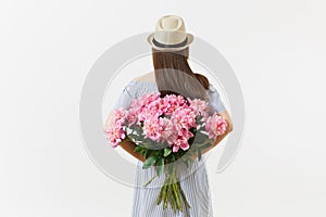 Young woman in blue dress, hat holding bouquet of beautiful pink peonies flowers behind her back isolated on white