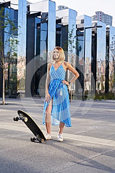 Young woman in blue dress with open shoulders stands with longboard or skate outdoor in a park.