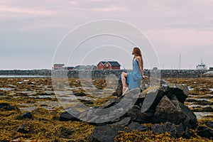 Young woman in blue dress sitting on stone among algae in tide