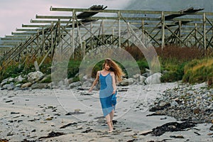 Young woman in blue dress walking on white sand, fish dryers on background