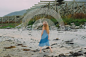 Young woman in blue dress walking on white sand, fish dryers on background