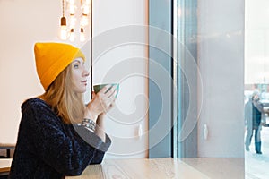 Young woman with blue eyes and blond hair in a yellow knitting hat is drinking coffee in a cafe.