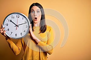 Young woman with blue eyes doing countdown holding big clock over yellow background scared in shock with a surprise face, afraid