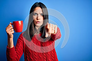 Young woman with blue eyes drinking cup of coffee standing over isolated background pointing with finger to the camera and to you,