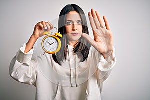 Young woman with blue eyes holding alarm clock standing over isolated white background with open hand doing stop sign with serious