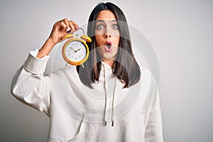 Young woman with blue eyes holding alarm clock standing over isolated white background scared in shock with a surprise face,