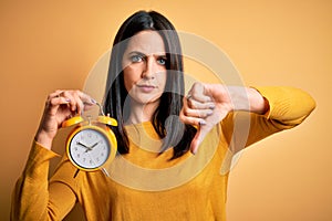 Young woman with blue eyes holding alarm clock standing over isolated yellow background with angry face, negative sign showing