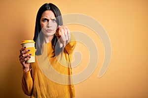 Young woman with blue eyes holding cup of coffee standing over yellow background pointing with finger to the camera and to you,
