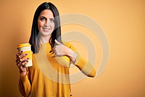 Young woman with blue eyes holding cup of coffee standing over yellow background with surprise face pointing finger to himself