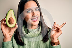 Young woman with blue eyes holding middle healthy avocado over isolated white background very happy pointing with hand and finger