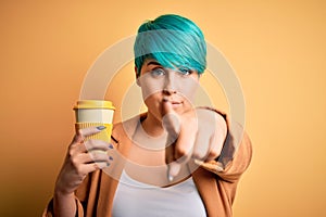 Young woman with blue fashion hair drinking a cup of coffee over yellow isolated background pointing with finger to the camera and