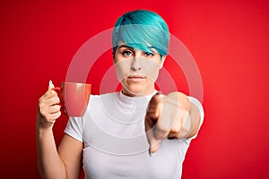 Young woman with blue fashion hair drinking a cup of coffee s over red isolated background pointing with finger to the camera and