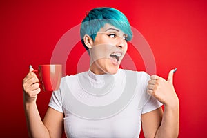 Young woman with blue fashion hair drinking a cup of coffee s over red isolated background pointing and showing with thumb up to