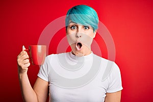 Young woman with blue fashion hair drinking a cup of coffee s over red isolated background scared in shock with a surprise face,