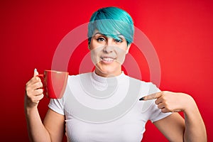 Young woman with blue fashion hair drinking a cup of coffee s over red isolated background with surprise face pointing finger to