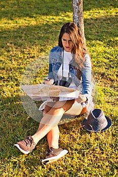 Young woman with blue hat, denim jacket and binoculars consulting a map and compass during a trip sitting on a park floor