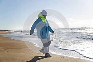 A young woman in a blue jacket runs away from the sea wave on a sunny day in winter. Rest and relaxation in nature