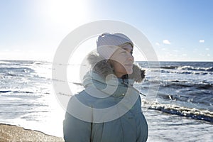 A young woman in a blue jacket walks along the seashore in the winter season on a sunny day. Active lifestyle and walks in nature
