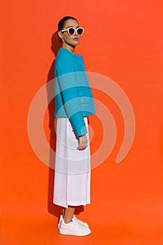 Young woman in blue jacket, white baggy pants and sneakers is standng. Side view