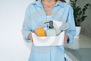 Young woman in blue shirt holding box of things for cleaning the kitchen at home