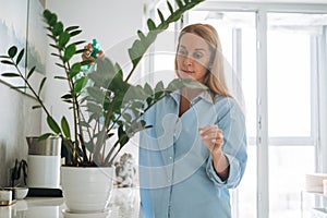Young woman in blue shirt with spray with water in hands takes care of houseplant in kitchen at home