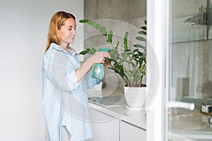 Young woman in blue shirt with spray with water in hands takes care of houseplant in kitchen at home