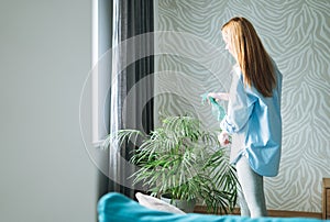 Young woman in blue shirt with spray with water in hands takes care of houseplant in living room at home